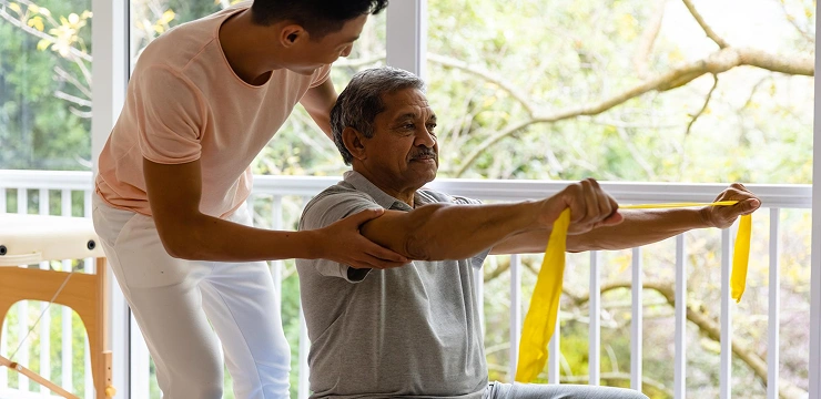 A therapist helps an older man exercise with a yellow resistance band.