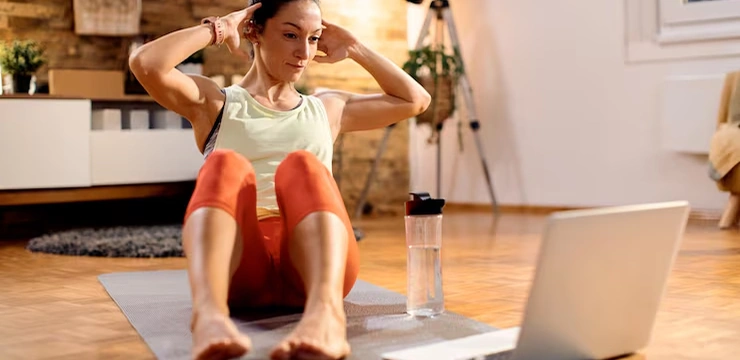 A woman does sit-ups at home while watching a workout on her laptop.
